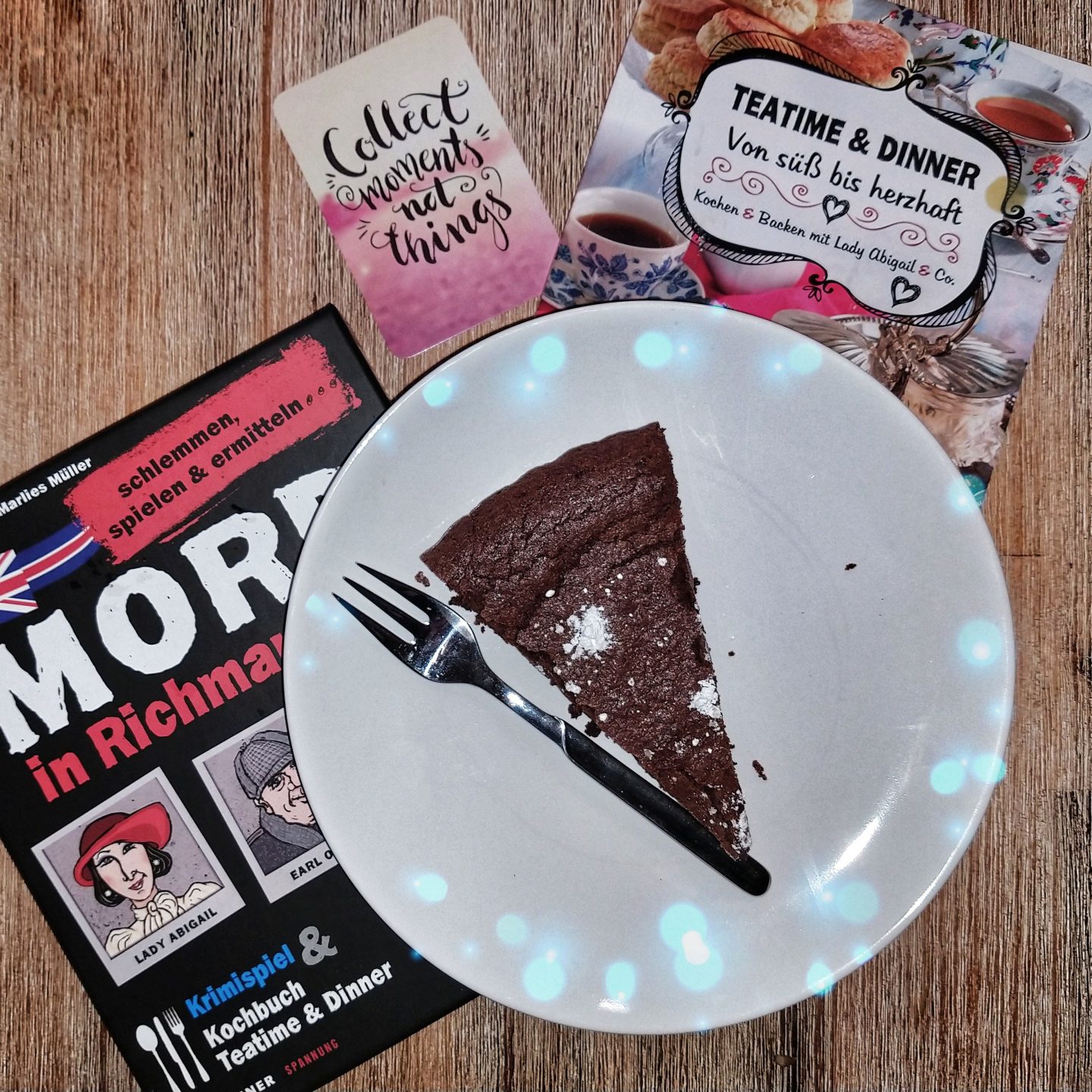 Die Verpackung des Krimi-Dinners Mord in Richman Hall neben einem Stück Schokoladen Tarte auf einem Teller und dem Kochbuch zum Spiel