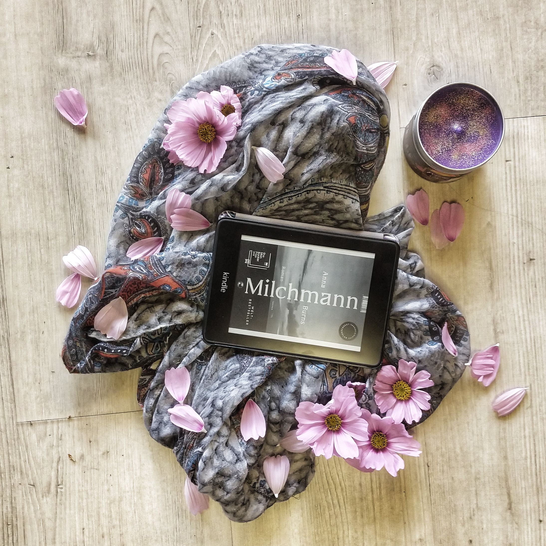Der Kindle zeigt das Cover von Milchmann in schwarz-weiß. Das Gerät liegt auf einem Schal auf einem hellem Holzboden, darum rosa Blüten und eine lila Kerze
